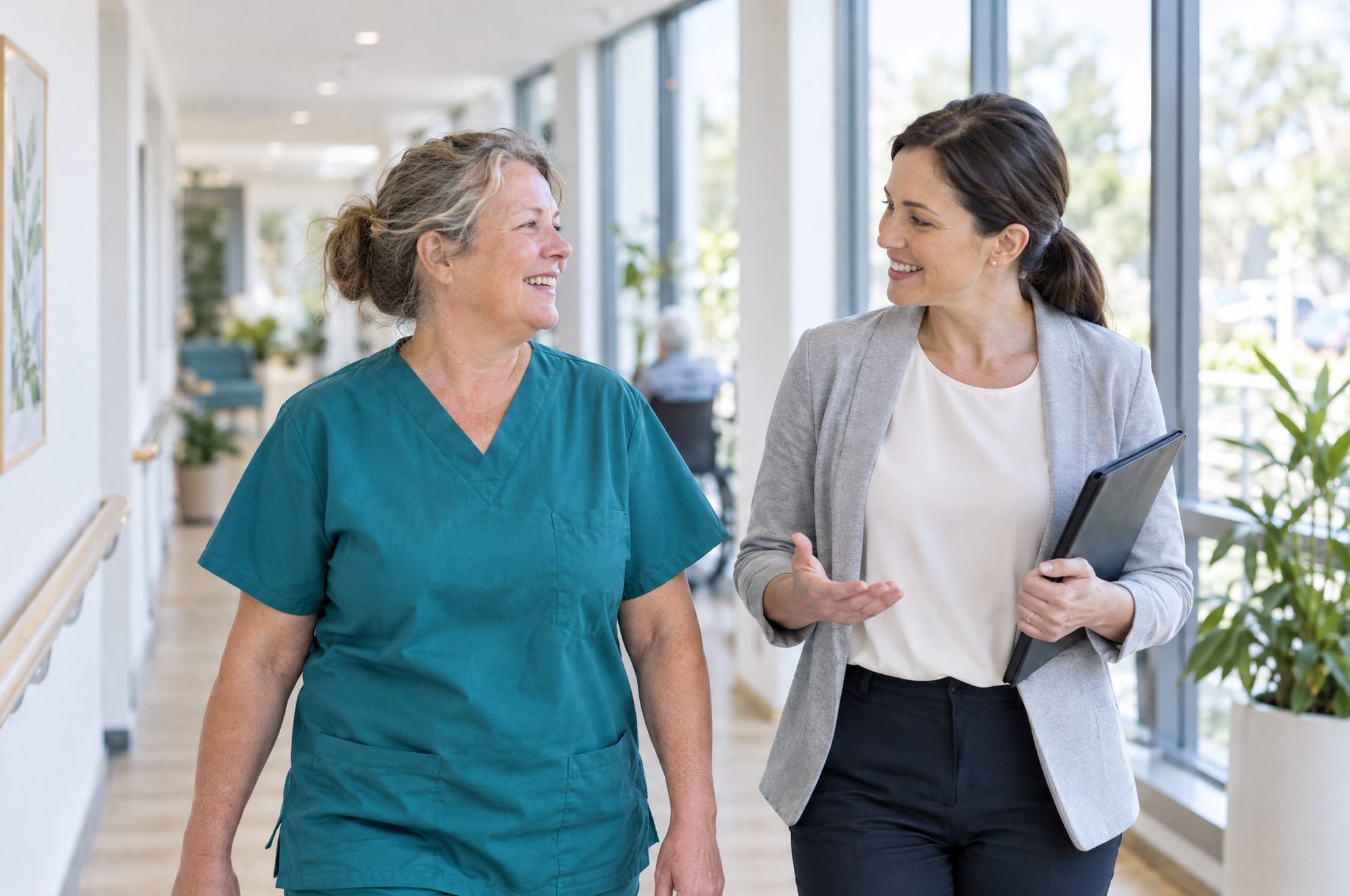 A care worker in uniform smiling while talking with a consultant in a bright corridor